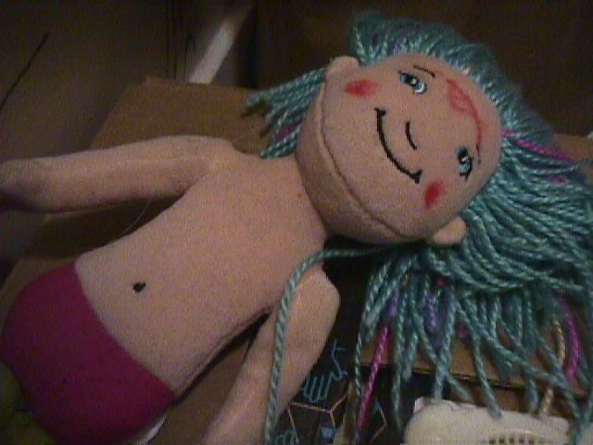 A doll with yarn hair laying o  a box, smiling upward 