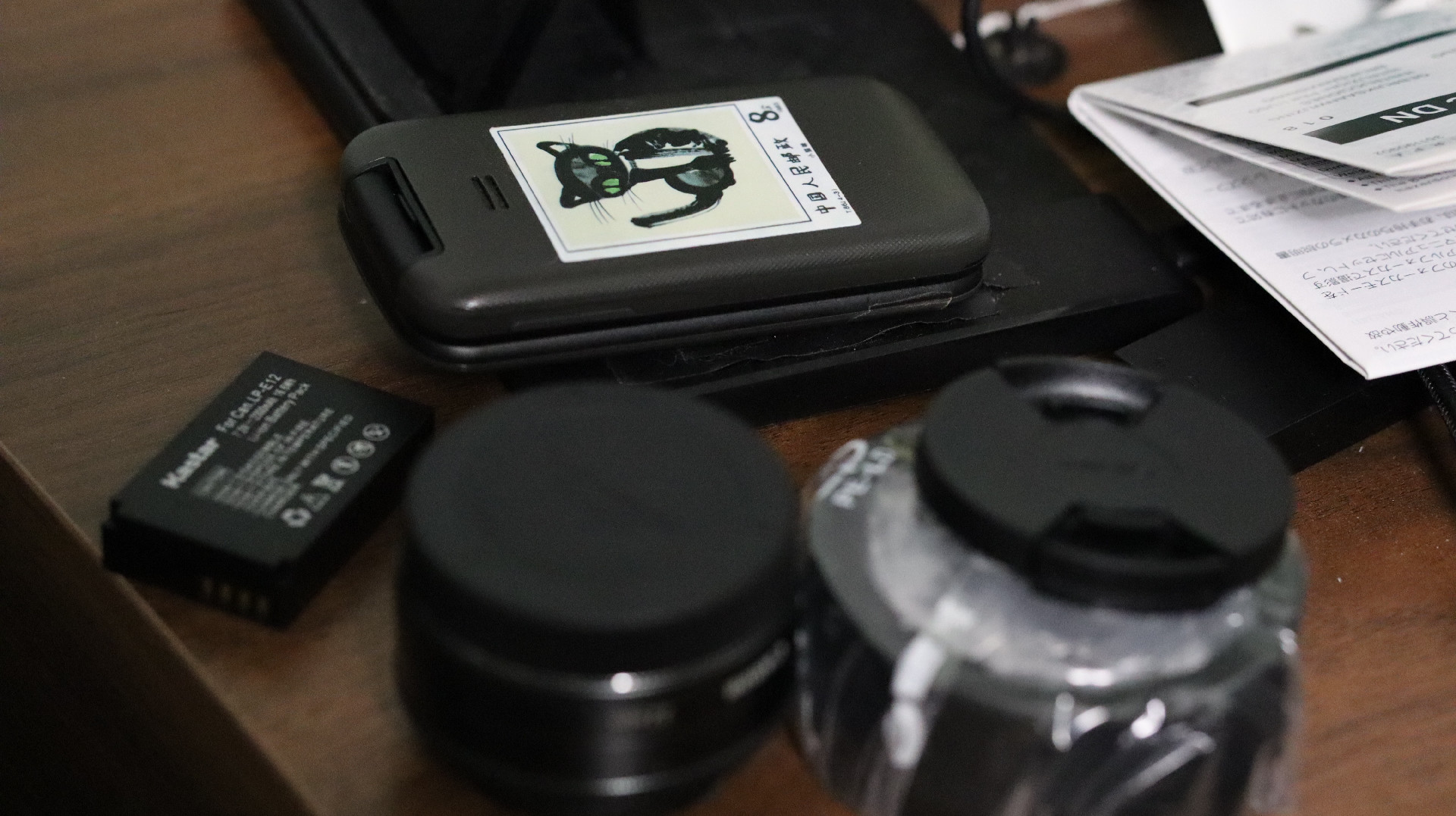 Items on a bedside table. Flip phone with a sticker of a black cat, two camera lenses and a camera battery.